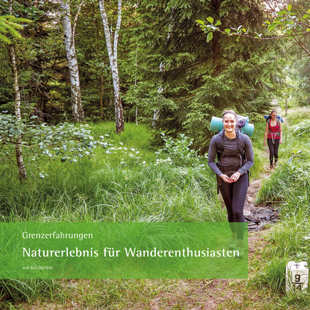 Zwei Wanderinnen mit Rucksäcken laufen auf einem schmalen Pfad durch den Wald.