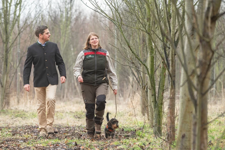 Ein Waldbesitzer läuft mit einer Försterin, die einen kleinen Hund mit sich führt, durch den Wald.