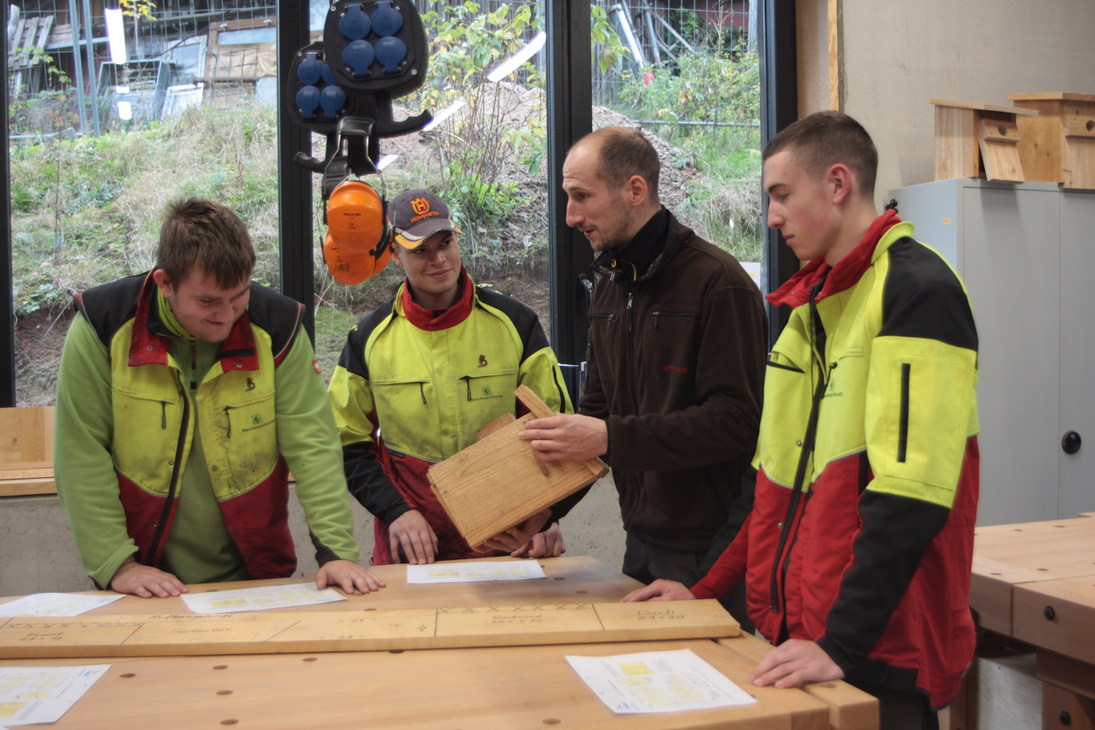 Drei Lehrlinge und ein Lehrmeister stehen an einem Tisch. Der Lehrmeister erklärt und die Lehrlinge hören zu.