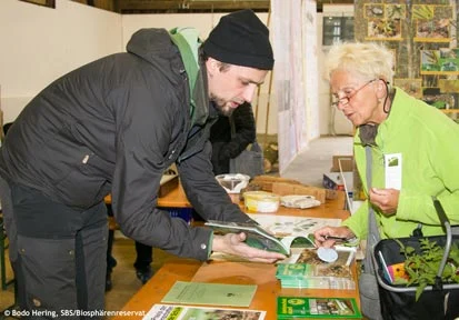 Ein Mitarbeiter am Stand zeigt einer &auml;lteren Dame eine Brosch&uuml;re
