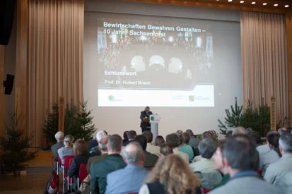 Blick &uuml;ber die Zuh&ouml;rer zur B&uuml;hne, wo der Vortragende vor einer Projektionswand referiert.