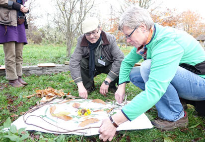 Zwei Workshop-Teilnehmer hocken auf einer Wiese vor einem großen Blatt Papier, auf den Zweige, Blätter und Blüten kreisförmig angeordnet sind.