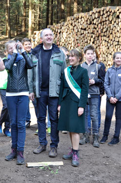 Eine Schülerin misst mit einem Baumhöhenmesser, der Minister und die Waldkönigin schauen lachend zu